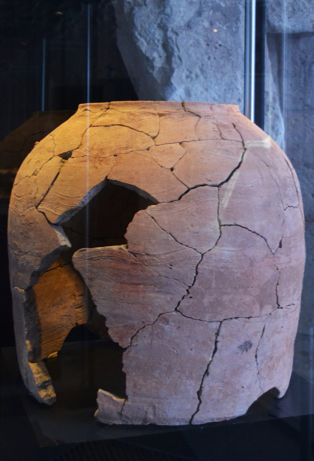 A cracked, tall ceramic tannur oven in a museum case