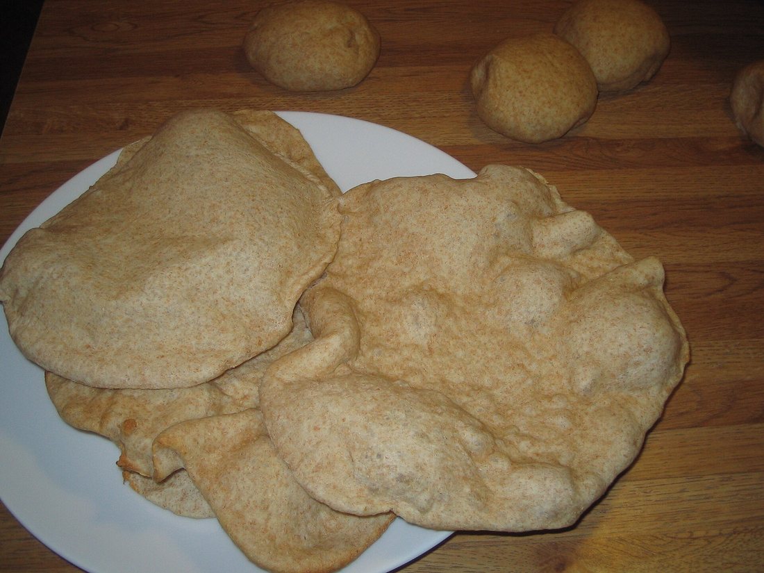 Several puffed homemade whole-wheat pitas on a plate, with unbaked dough balls behind