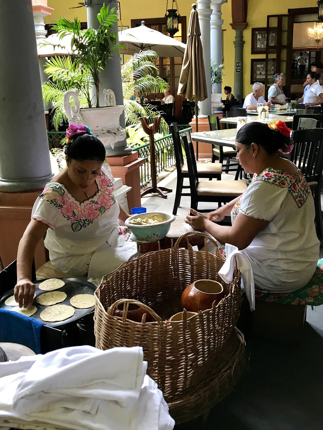 Two women in white embroidered huipiles making corn tortillas by hand at a comal in a restaurant courtyard