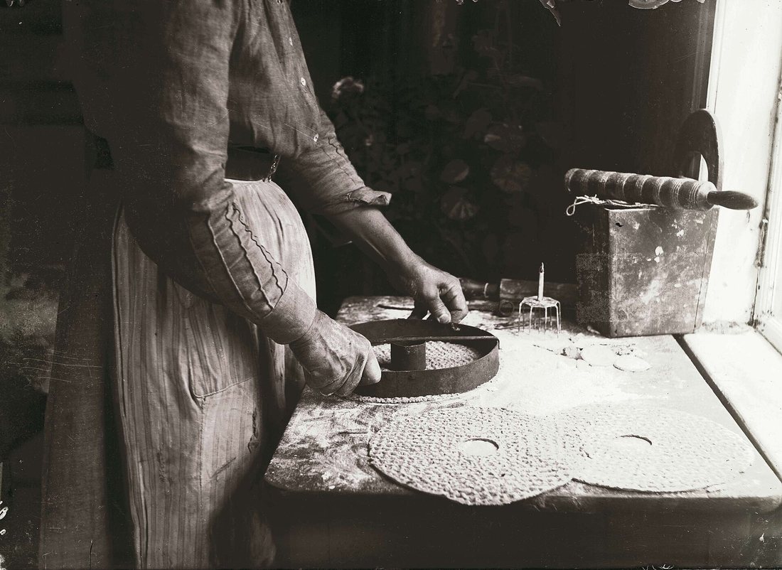 An old black-and-white photograph of a woman at a floured table shaping round crispbreads with holes in the centre