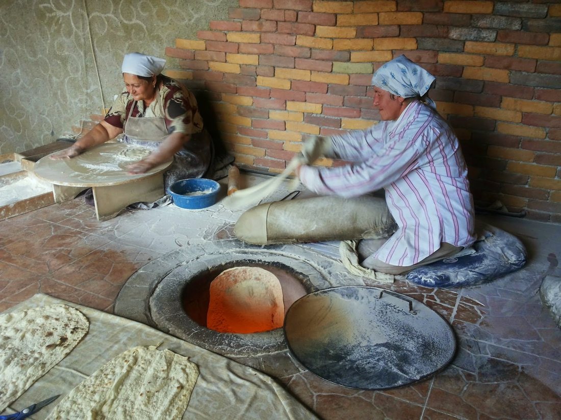 Two women in headscarves making lavash in Armenia: one stretches dough on the floor, the other kneels beside a floor-set tonir with its glowing mouth open