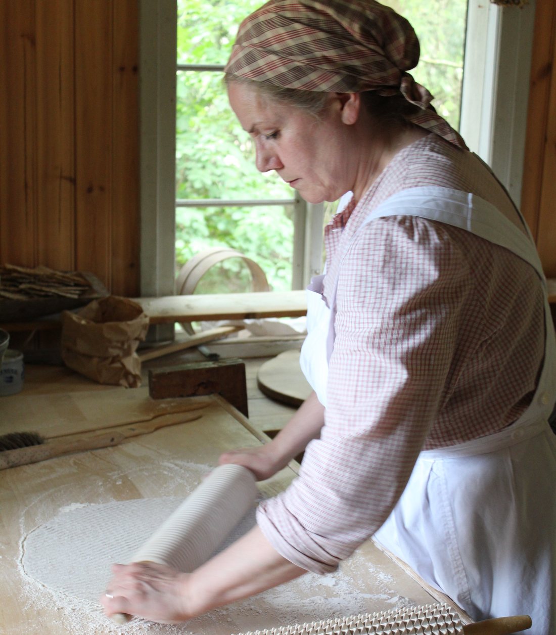A woman in historical dress and headscarf rolling a large thin sheet of dough with a rolling pin at a wooden table