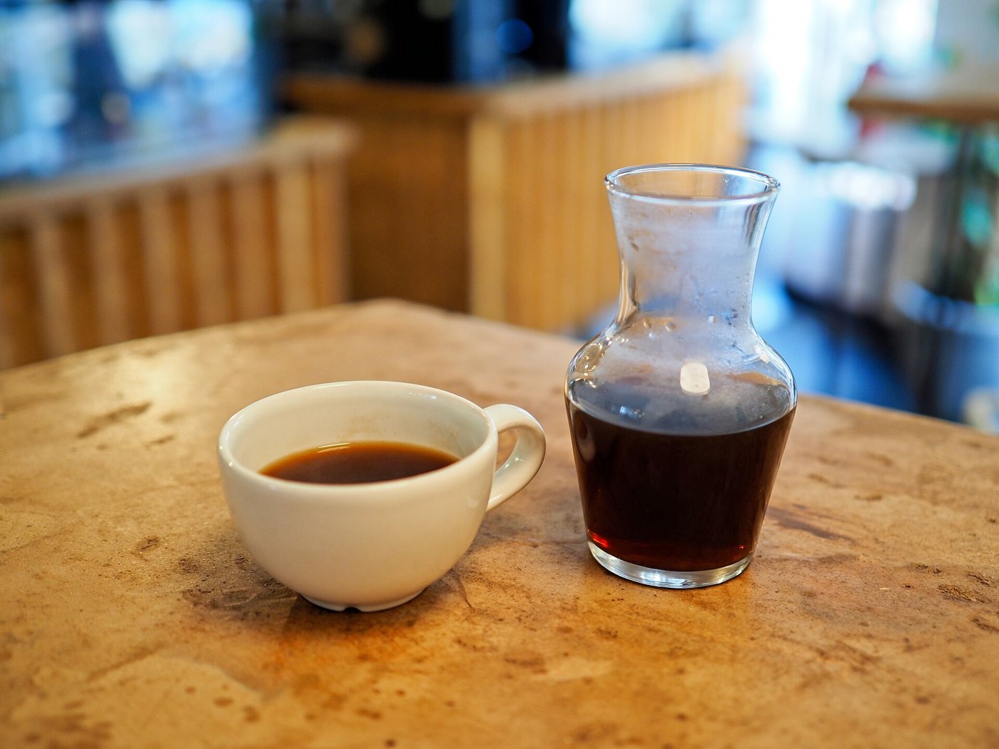 A white cup of black coffee and a small glass carafe half full of coffee on a worn café table