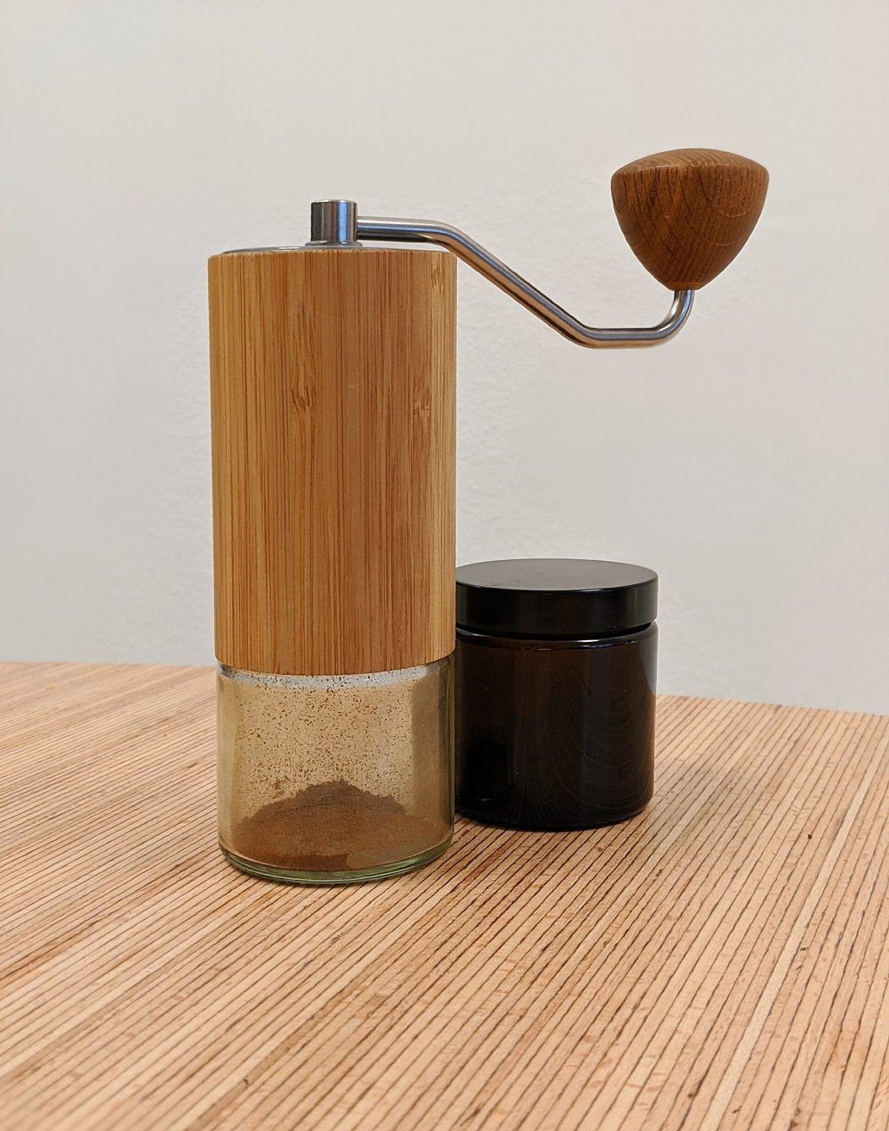 A hand coffee grinder with a wood-veneered body, a steel crank and a wooden knob, standing on its glass grounds jar beside a spare amber jar