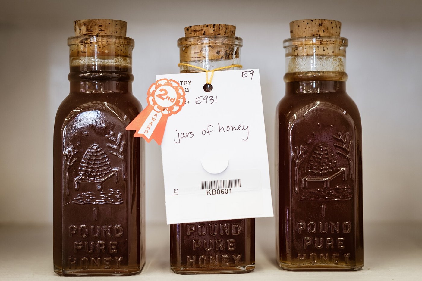 Three square glass jars of dark honey with cork stoppers and a beehive moulded into the glass, one with a second-place rosette and a handwritten entry tag reading jars of honey