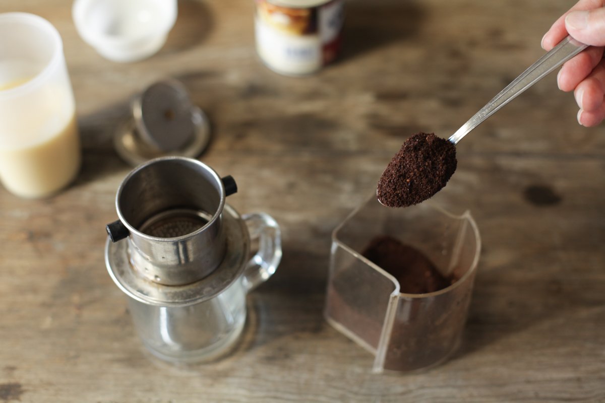 A spoonful of ground coffee held over a steel phin filter that sits on a glass mug, with a clear container of grounds and a glass of condensed milk on a wooden table