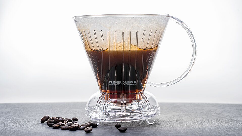 A clear plastic Clever dripper full of steeping coffee standing on a counter with a few beans beside it
