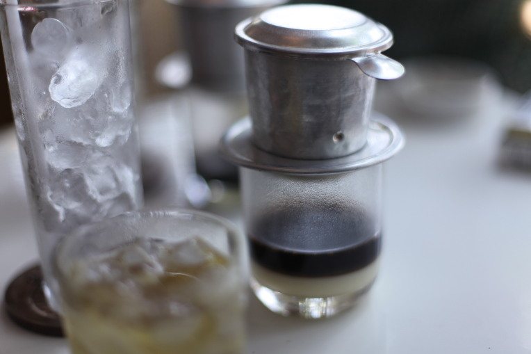 A steel phin filter dripping coffee into a glass over a white layer of condensed milk, with a tall glass of ice beside it