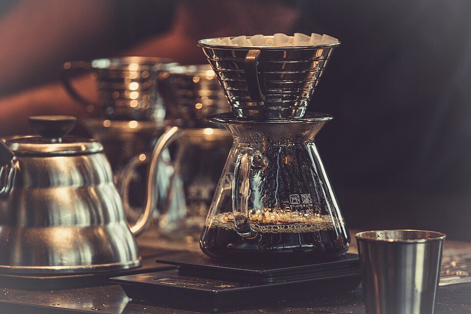 A glass pour-over brewer with a fluted paper filter of wet grounds standing on a scale, steel kettles beside it
