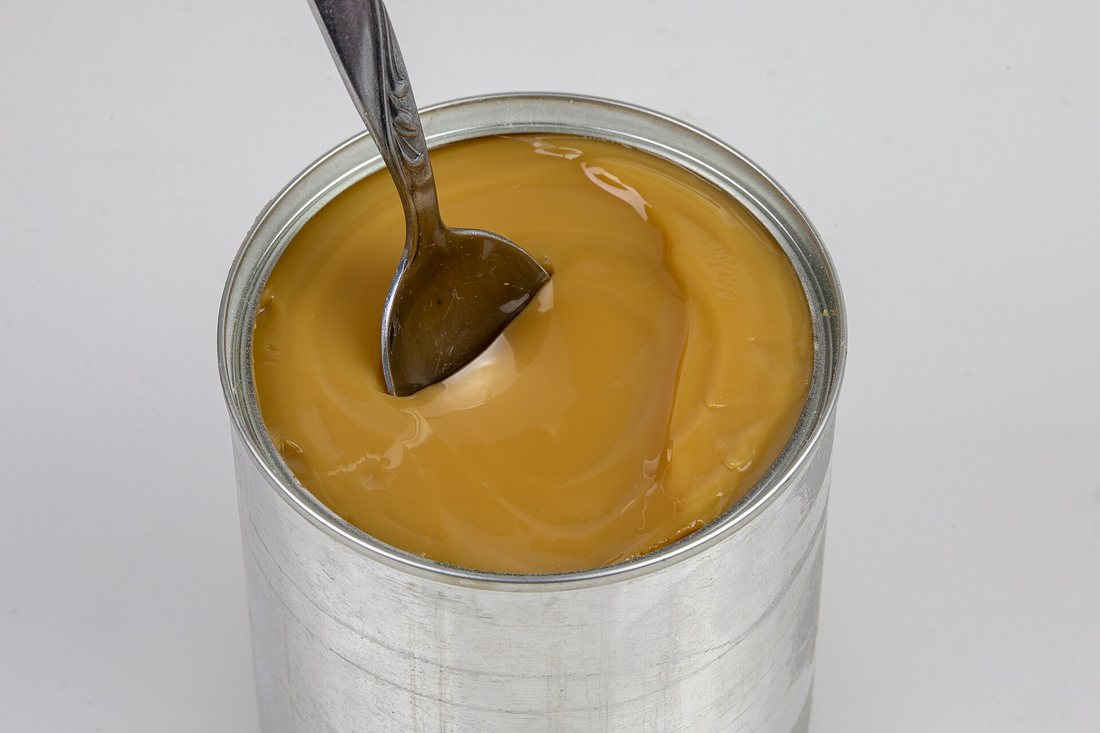 An opened can of caramelized condensed milk, tan and thick, with a spoon in it