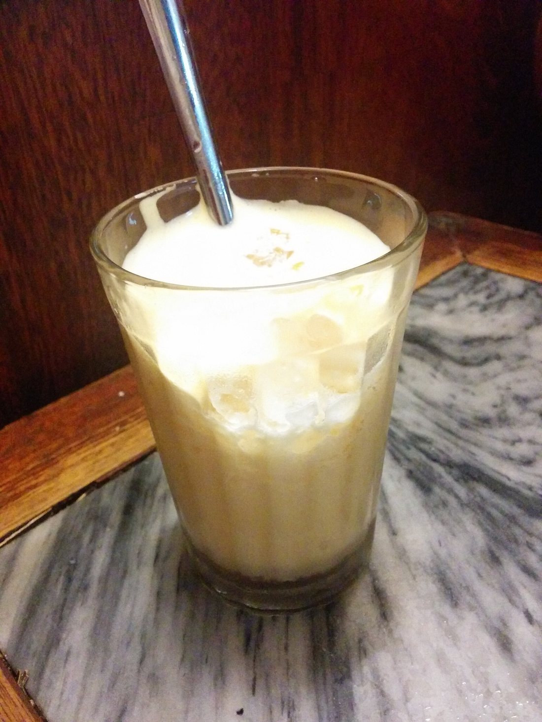 A glass of egg coffee topped with a thick pale foam, a metal straw in it