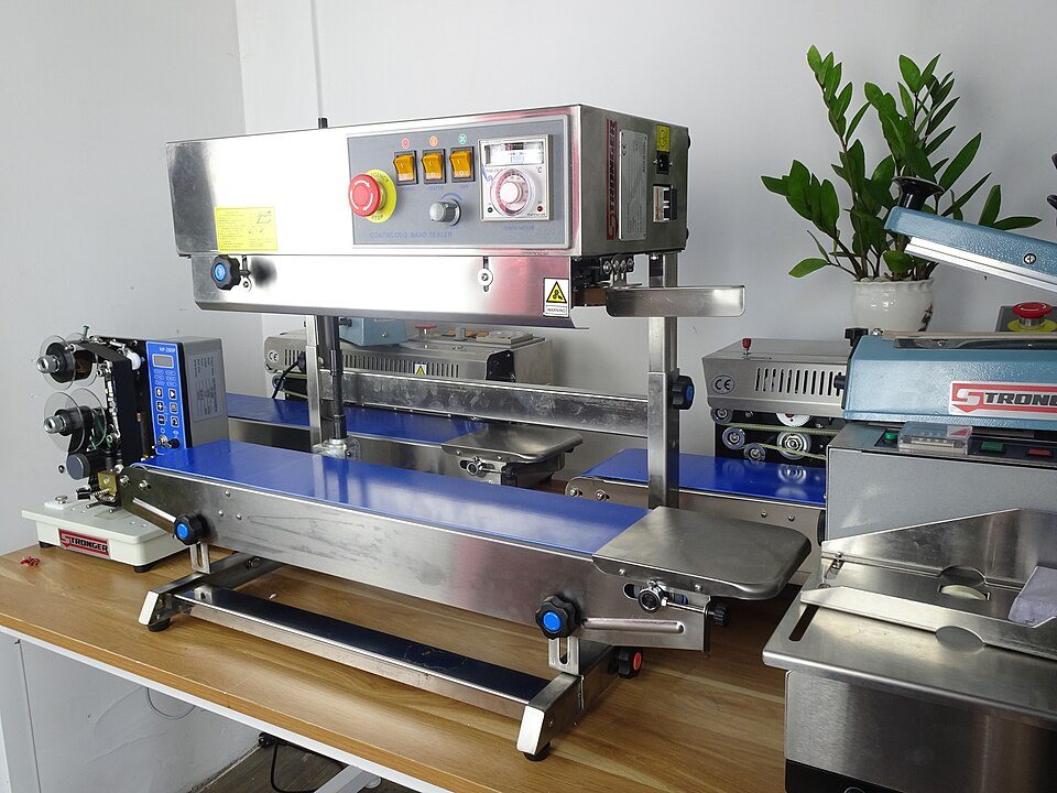 A stainless continuous band sealer on a bench: a conveyor belt under a sealing head with a control panel