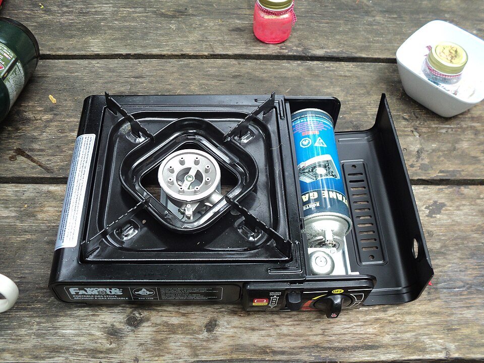 A single-burner portable gas stove on a wooden table, its side cover open to show the fuel canister