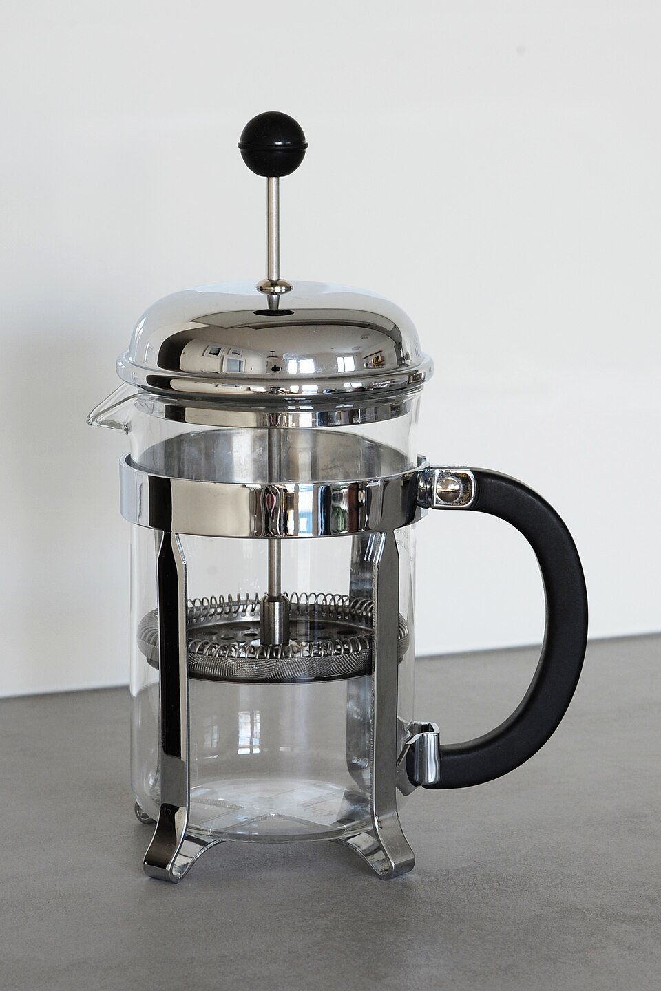 An empty glass French press with a chrome frame and a black handle and knob, on a grey counter