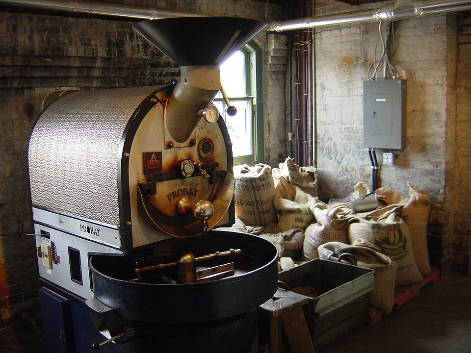 A drum roaster with a perforated steel housing and a cooling tray in a brick-walled room, sacks of green coffee beside it