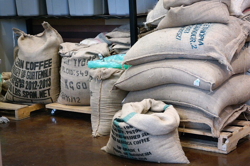 Burlap sacks of green coffee from several origins stacked on pallets in a roastery, one sack open at the top