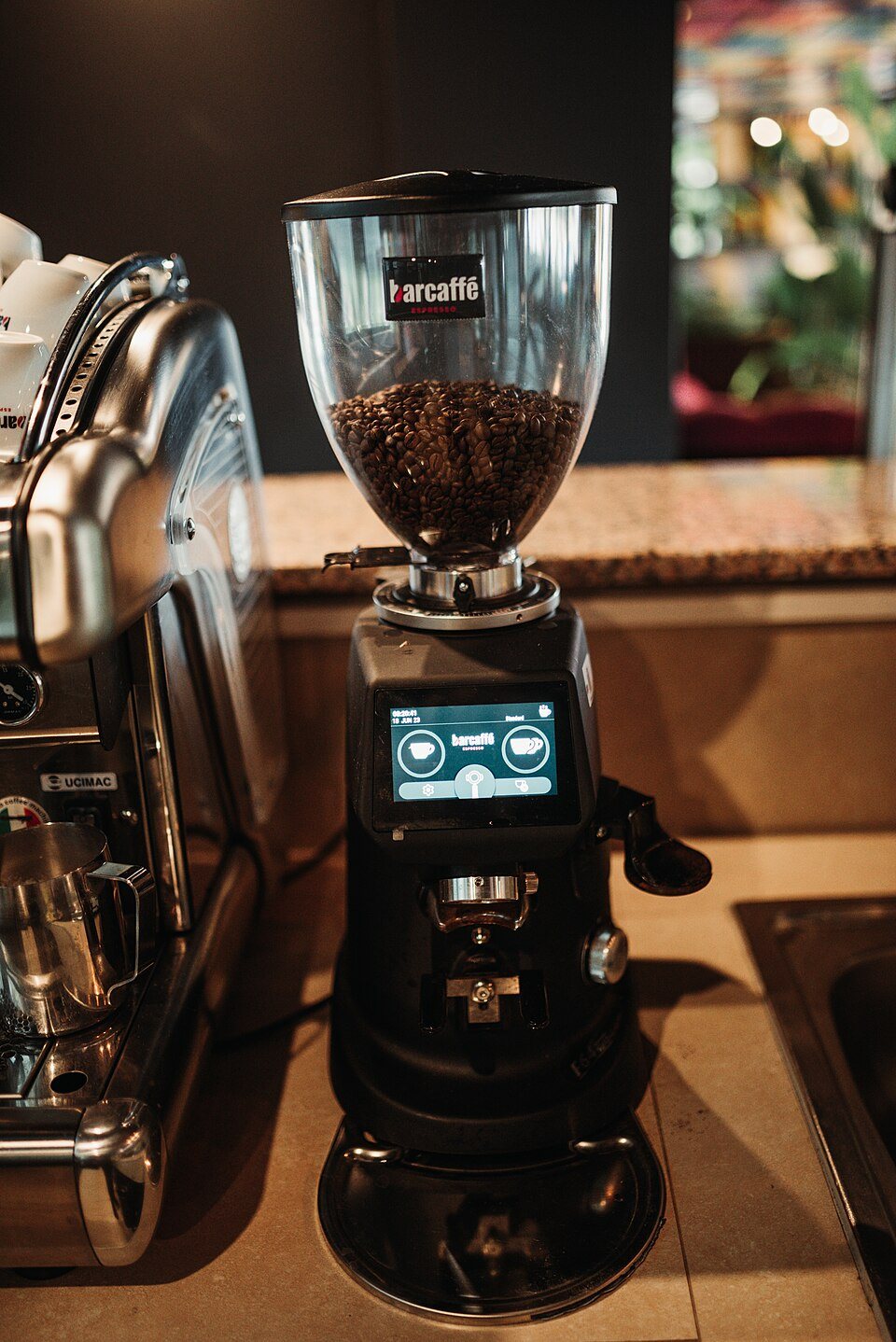 A tall café grinder with a hopper of beans and a touchscreen, standing beside an espresso machine