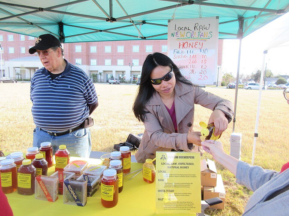 A vendor under a canopy serves a customer at a table of honey jars; a hand-lettered sign reads Local Raw New Orleans Honey with a list of sizes and prices