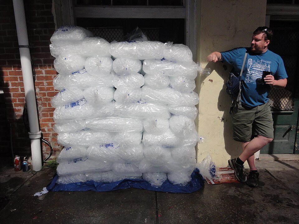 A man leans against a wall of stacked bags of ice taller than he is