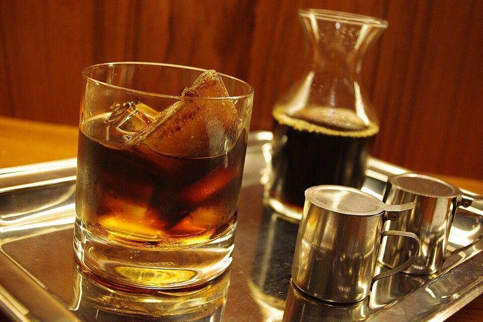 A short glass of dark coffee over large pieces of ice on a tray, with a small carafe and two steel pitchers