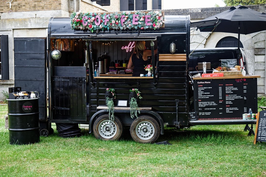 A black converted horse-box trailer on a lawn, its side hatch open as a coffee counter, a flowered sign reading COFFEE on the roof, airpots on the counter and a menu board at the tow-bar end