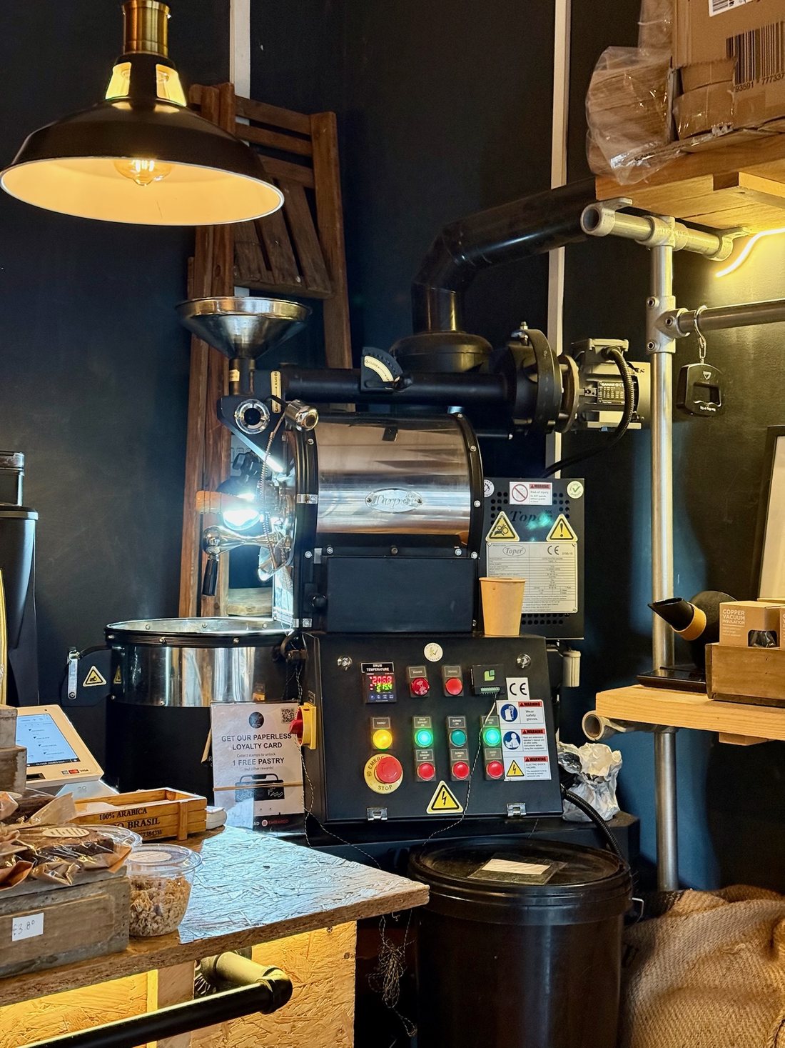 A small drum coffee roaster with a control panel and cooling tray in the corner of a shop