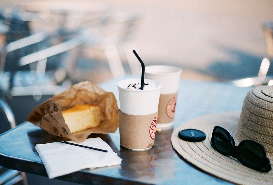 Two lidded paper coffee cups with sleeves on a café table beside a pastry in a paper bag