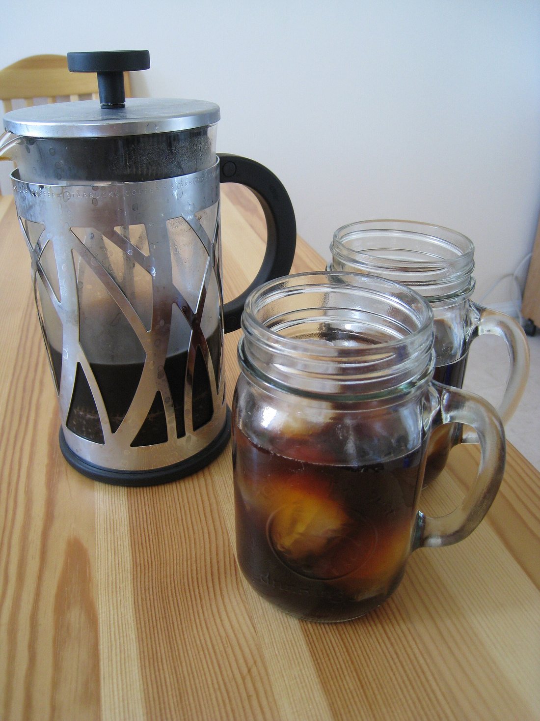 A glass French press in a steel frame beside two jar mugs of iced coffee on a wooden table