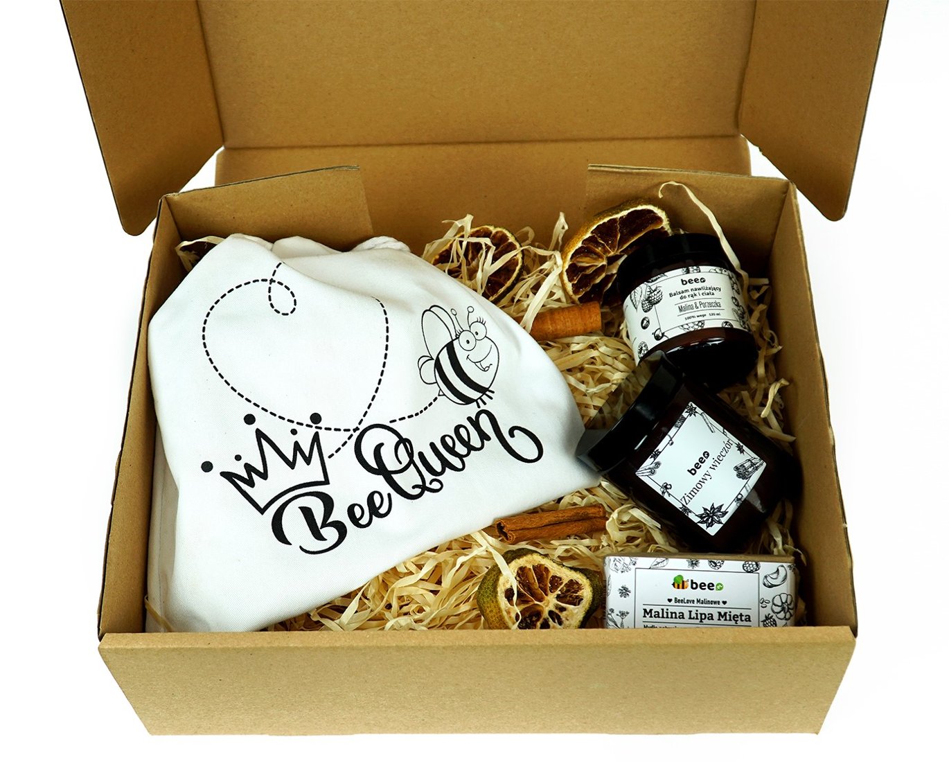 An open kraft cardboard box packed with pale wood wool, holding a white cloth bag printed with a cartoon bee and the words Bee Queen, two dark jars with white labels, a small printed carton, dried citrus slices and cinnamon sticks