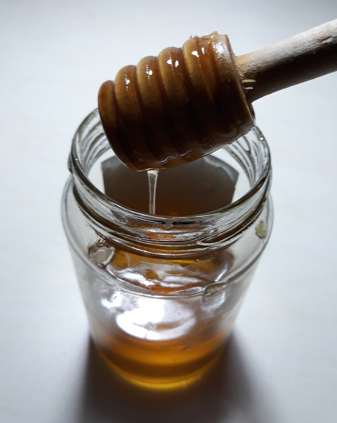 A wooden honey dipper lifted from a small glass jar, honey running from it