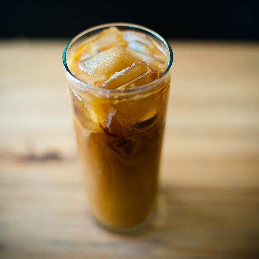 A tall glass of iced coffee with milk swirling through it, on a wooden counter