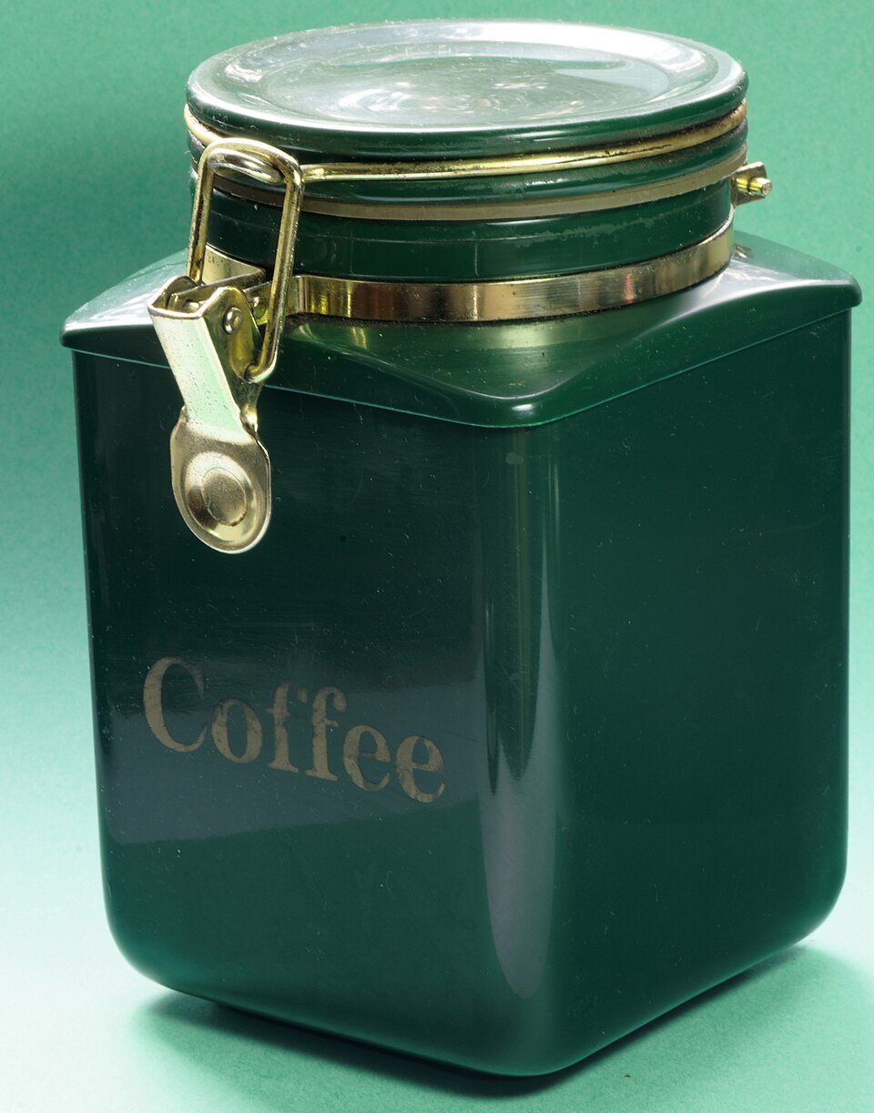 A square dark green storage tin with the word Coffee in gold on the front and a brass clip holding down a round lid
