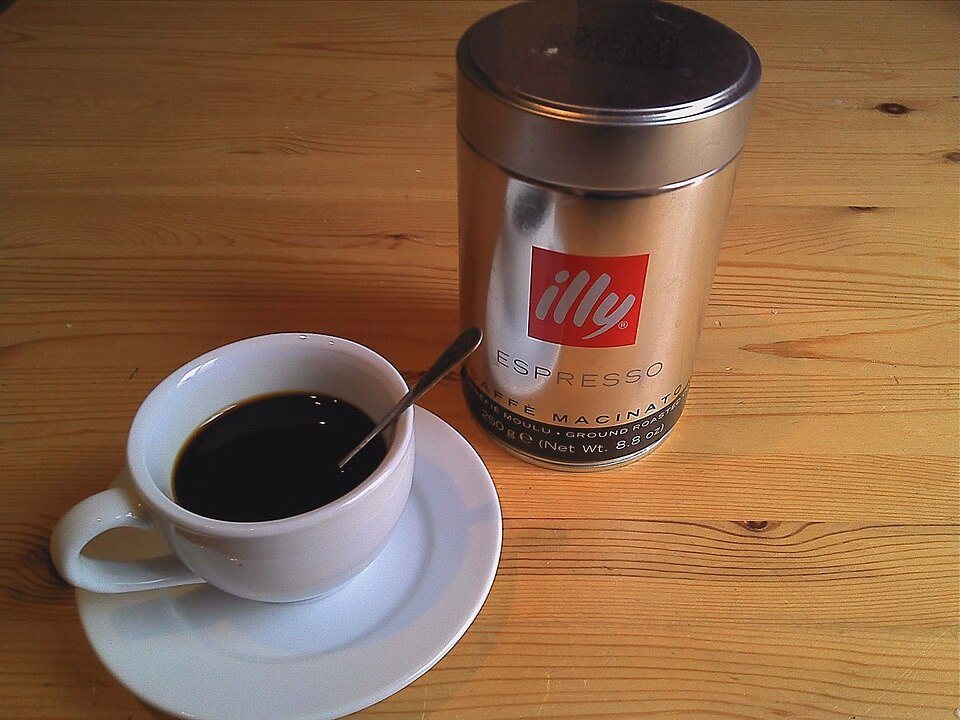 A silver Illy espresso can with its red logo standing on a wooden table beside a white cup of black coffee