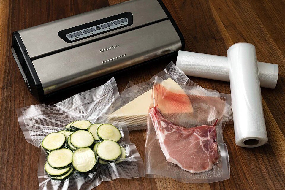 A countertop vacuum sealer on a wooden table with rolls of bag film and three sealed pouches holding sliced courgette, cheese and a pork chop