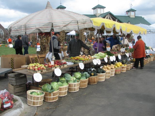 A long market table under striped umbrellas, with bushel baskets of cabbages, squash and apples and round price signs along the front