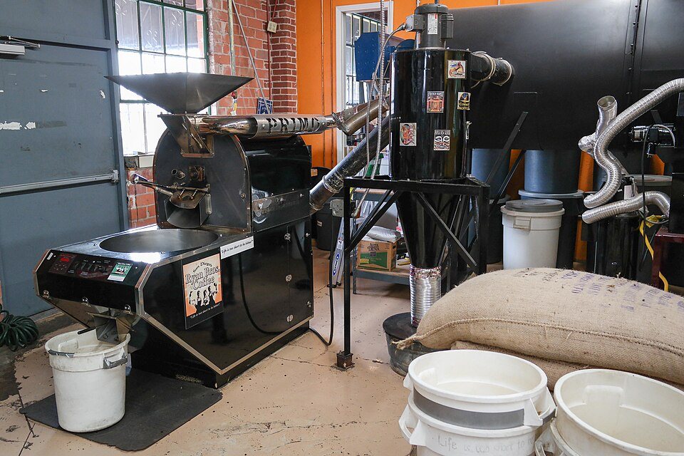 A black drum roaster in a workshop with a metal cyclone and ducting behind it and a sack of green coffee in front