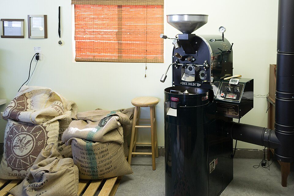 A small black drum roaster on a pedestal in the corner of a room, burlap sacks of green coffee stacked beside it