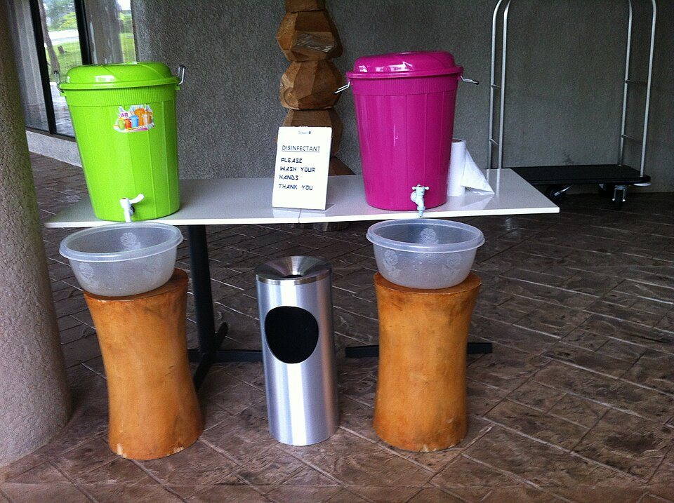 Two lidded plastic buckets with taps on a table, each above a basin on a stool, with a sign asking people to wash their hands