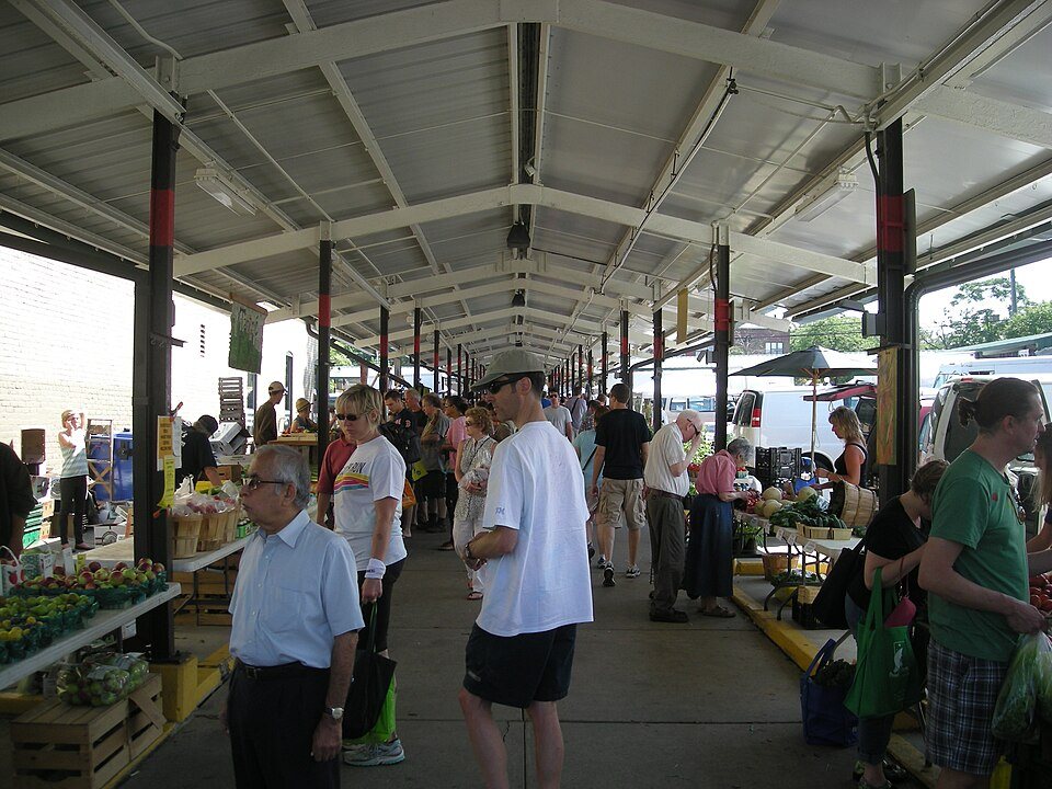 Shoppers walking down the aisle of a long open-sided market shed lined with produce tables