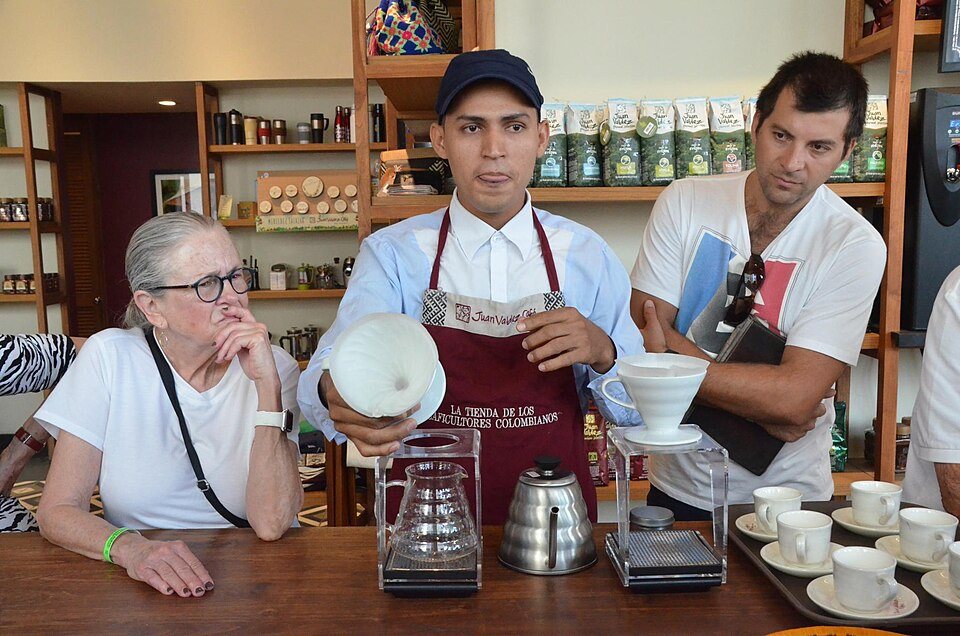 A barista holds up a white pour-over cone to show a group seated around a table of small cups