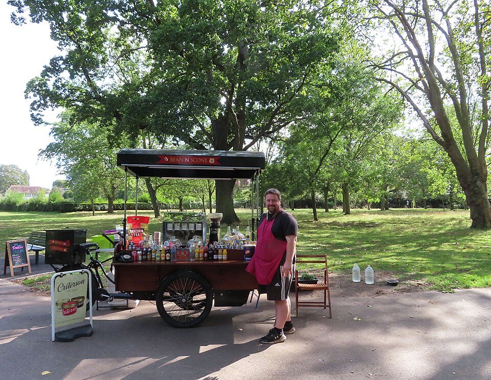 A man in an apron stands beside a coffee cart built on a cargo tricycle in a park, bottles of syrup lined up on its counter