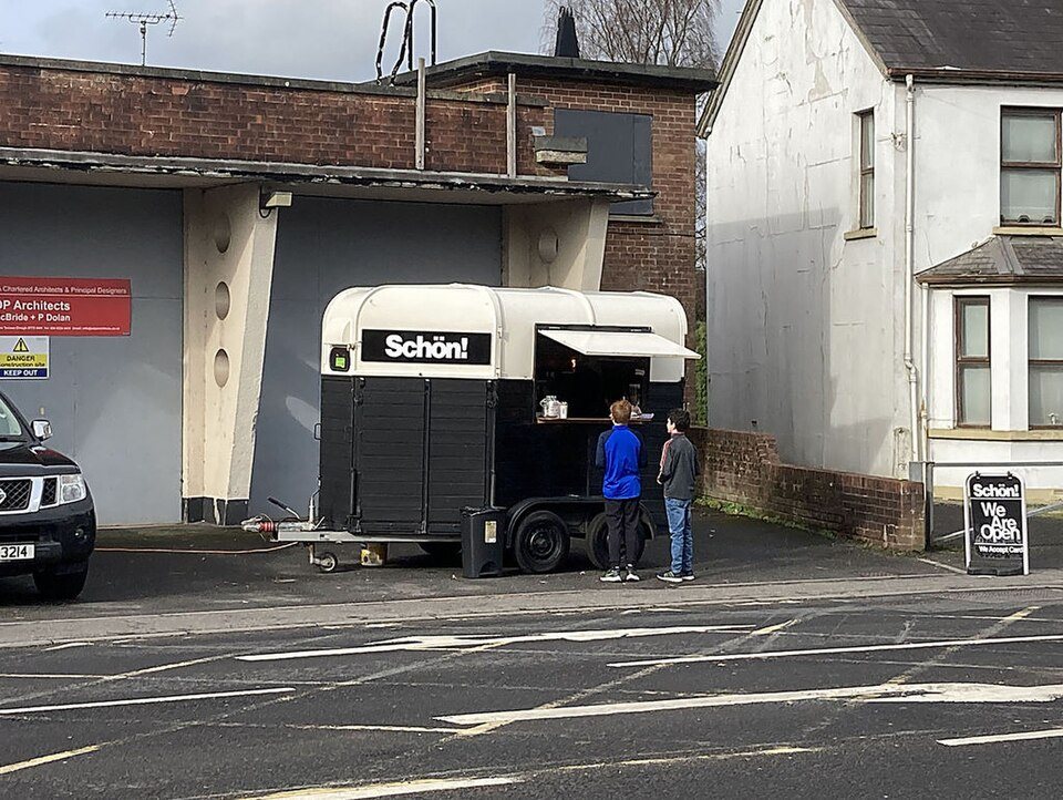 A black and cream trailer with a serving hatch and awning parked in a small car park, two customers at the window
