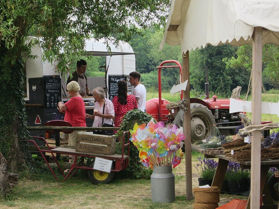 A small white trailer with a serving window among market stalls, flowers and a tractor on a farm