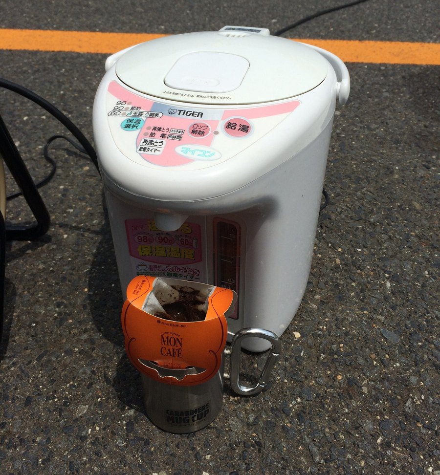 A white electric water boiler standing on asphalt with its power cord trailing away, and in front of it a steel mug with a paper drip-coffee filter hung on its rim