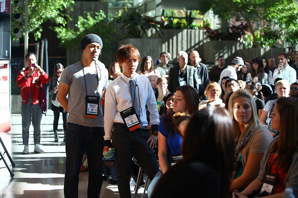 Two men wearing lanyards stand in front of a seated crowd at an indoor event