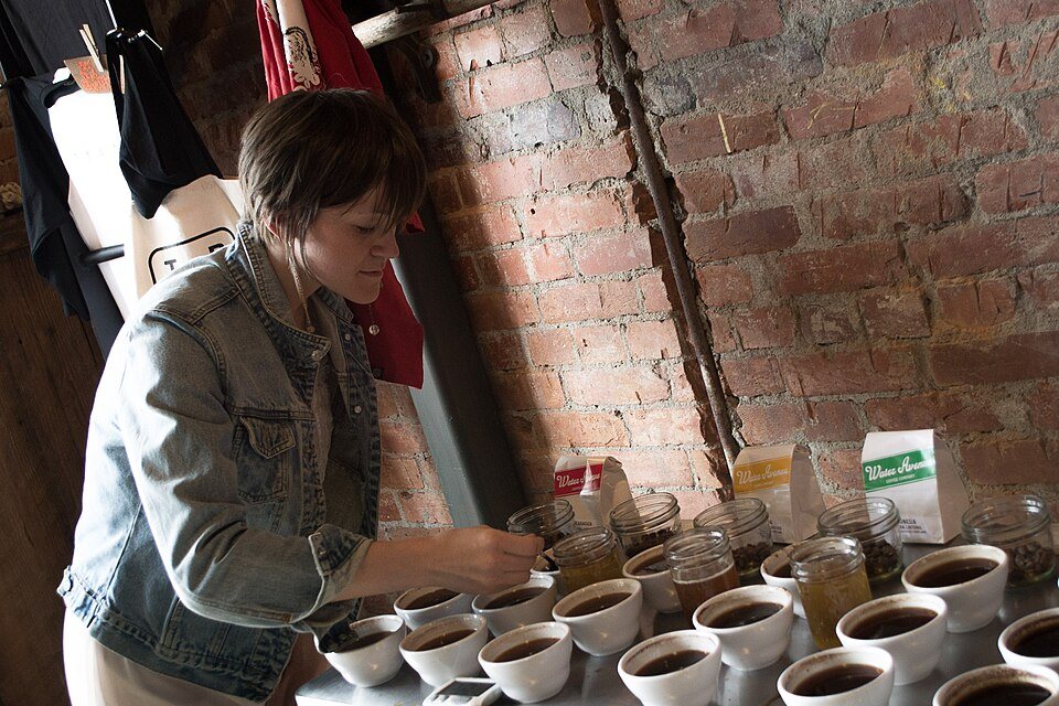 A woman dips a spoon into one of a dozen white bowls of coffee set out on a steel counter with mason jars and a bag of beans, against a brick wall