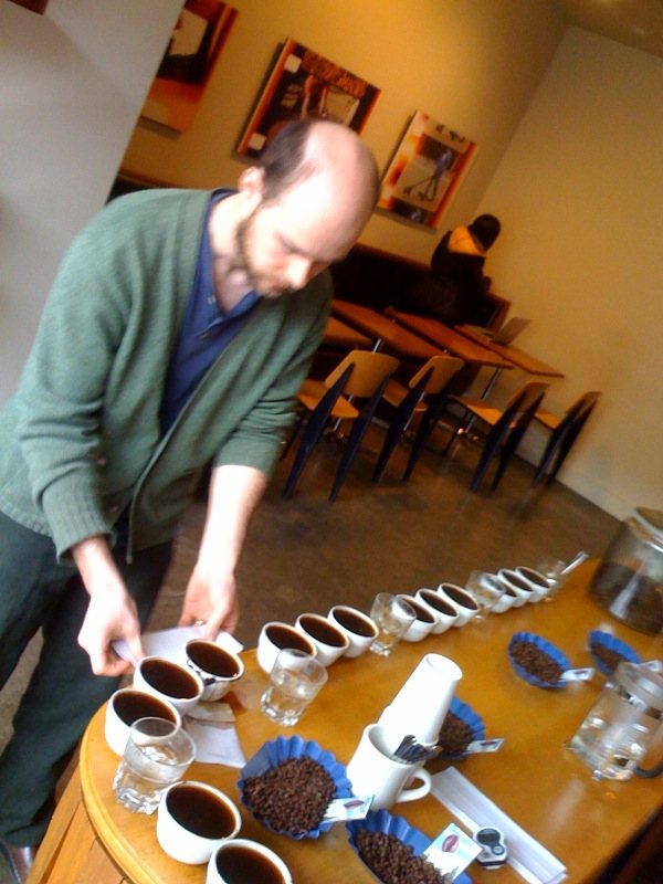 A man leans over a table set with a line of white cups of coffee, glasses of water and paper trays of roasted beans, a spoon in his hand