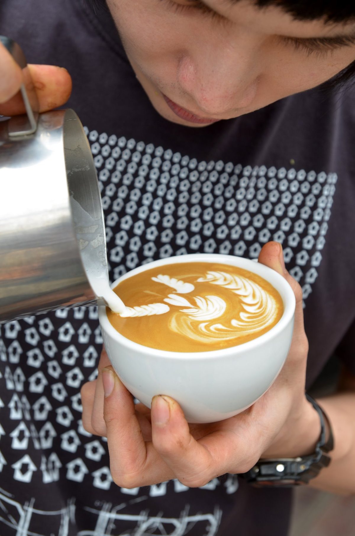 A pitcher of glossy, paint-textured microfoam being poured into a cortado, the foam folding into a fine ring