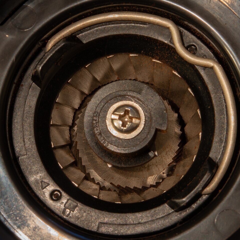 Looking straight down into a conical burr grinder with the hopper off: a toothed steel ring around a ridged cone held by a screw, with a film of fine brown coffee dust on the metal and the plastic around it