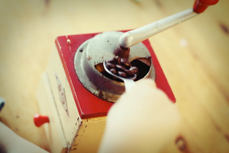 A red-topped wooden box mill seen from above, a spoonful of roasted beans being tipped into its small open hopper beside the crank